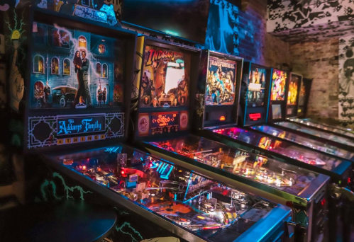 A row of colorful pinball machines featuring various themes in a dimly lit arcade.
