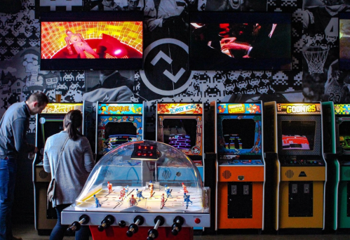 A retro arcade with colorful game machines, foosball table, and screens displaying games in a lively atmosphere.