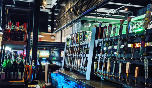 A vibrant bar with a variety of beer taps, colorful bottles, and arcade games in the background.