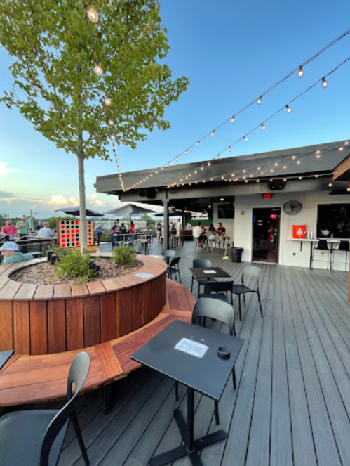 Outdoor dining area with wooden tables, string lights, and a central tree planter, creating a cozy atmosphere.