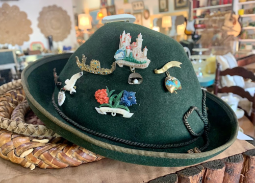 A green felt hat adorned with various colorful pins and brooches, displayed in a vintage shop setting.