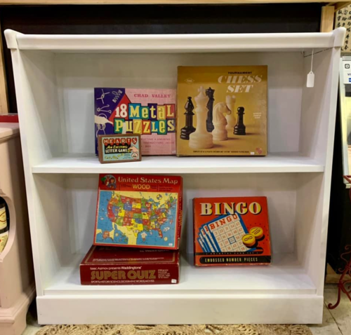 A white shelf displaying various board games and puzzles, including chess, bingo, and a United States map quiz.