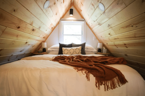 Cozy attic bedroom with wooden walls, a large bed, and decorative pillows, featuring warm lighting and a throw blanket.