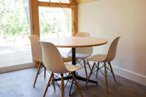 A round wooden table with four white chairs in a bright, minimalist room with large windows.