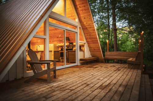 A cozy A-frame cabin with a wooden deck, surrounded by trees and warm interior lighting.
