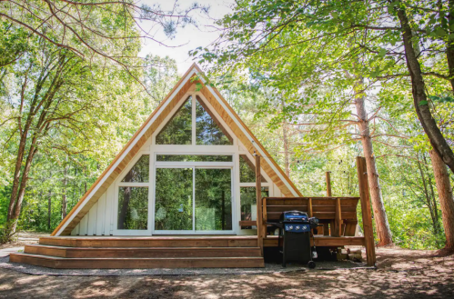 A modern A-frame cabin surrounded by trees, featuring large windows and a wooden deck with a grill.