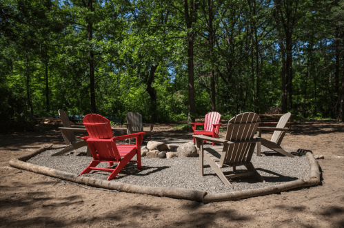 A circular fire pit area with red and gray Adirondack chairs surrounded by trees in a wooded setting.