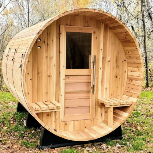 A wooden barrel-shaped sauna with a glass door, set in a forested area with autumn foliage.