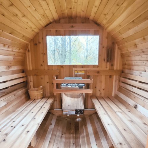 Interior of a wooden sauna with benches, a stove, and a window overlooking trees. Warm, inviting atmosphere.