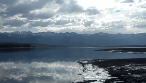 A serene lake reflects mountains under a cloudy sky, with a calm shoreline and distant snow-capped peaks.