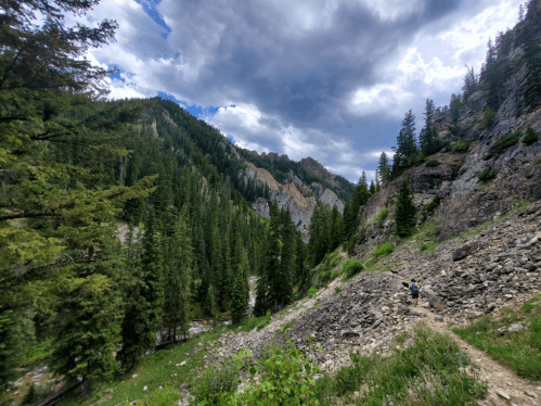 A hiker walks along a rocky trail surrounded by tall trees and mountains under a cloudy sky.