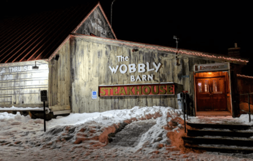 A rustic wooden building at night, featuring a sign that reads "The Wobbly Barn Steakhouse" with snow in front.
