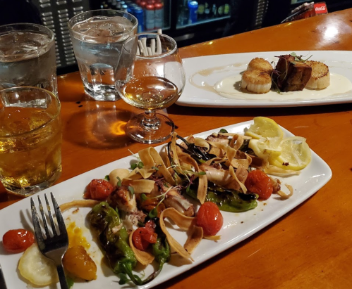 A table with two plates of gourmet food, glasses of water, whiskey, and a dessert, set in a cozy dining atmosphere.