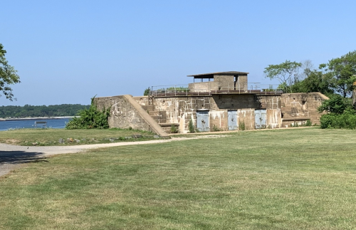 A historic stone fortification with a lookout tower, surrounded by grass and overlooking a calm body of water.