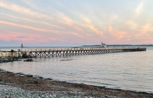 A wooden pier extends into calm waters under a pastel sunset sky, with a distant lighthouse visible.