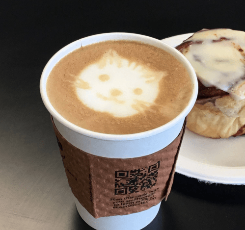 A coffee cup with a cat face latte art, next to a cinnamon roll on a plate.