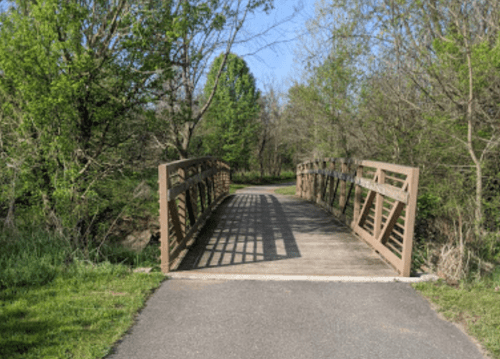 A wooden bridge spans a path surrounded by lush green trees and grass under a clear blue sky.