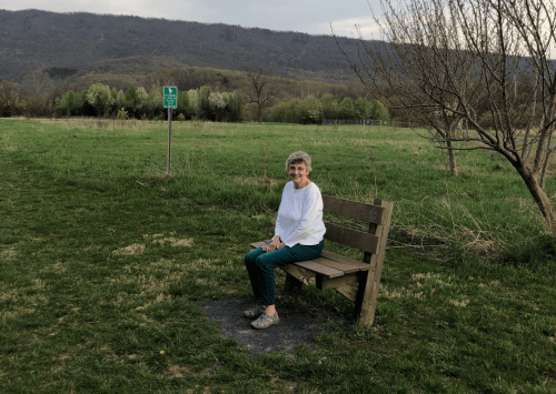 An older woman sits on a wooden bench in a grassy field with mountains in the background and a sign nearby.