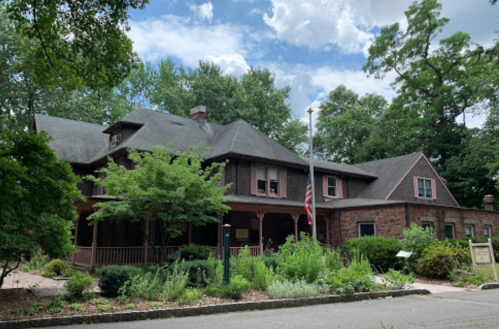 A large, historic house surrounded by greenery, featuring a porch and an American flag. Bright, cloudy sky above.