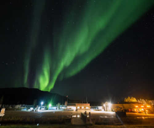 A vibrant green aurora borealis dances over a dark landscape, illuminating nearby buildings under a starry night sky.