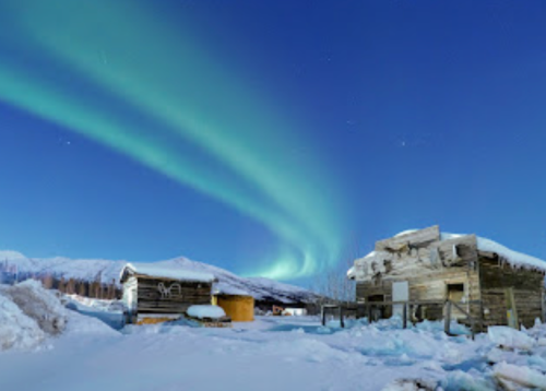 A snowy landscape at night with vibrant green auroras dancing in the sky above rustic wooden buildings.