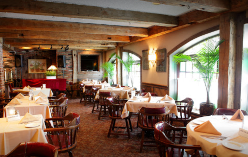 Cozy restaurant interior with wooden beams, tables set for dining, and large windows letting in natural light.