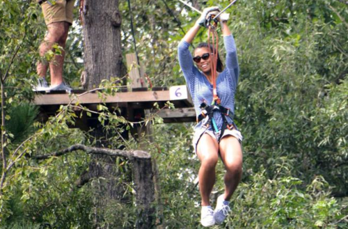 A woman smiles while zip-lining through a forest, wearing safety gear and enjoying the adventure.