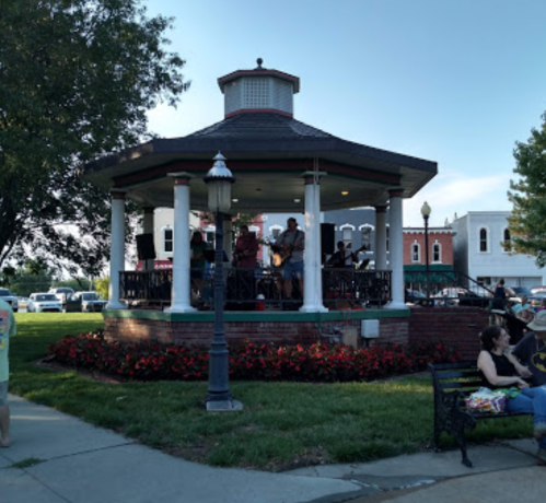 A band performs on a gazebo stage in a park, with people enjoying the music nearby. Green grass and flowers surround the area.