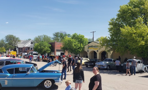 A lively street scene with classic cars, people mingling, and a building labeled "jamboree" under a clear blue sky.