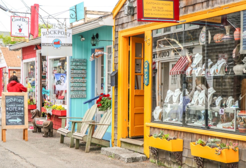 Colorful storefronts line a quaint street, featuring shops, signs, and decorative displays in a charming coastal town.