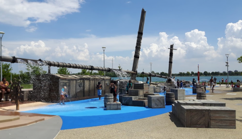 A water play area with wooden structures, children playing, and a scenic waterfront under a blue sky.