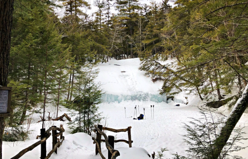 A snowy landscape with a frozen waterfall, surrounded by trees, and a person sitting on the ground with ski poles.