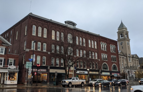 Historic brick buildings line a street, with shops on the ground floor and a clock tower in the background.