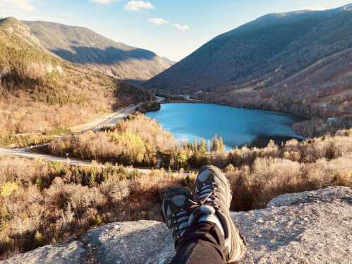 A pair of hiking shoes resting on a rock, overlooking a serene lake surrounded by mountains and autumn foliage.