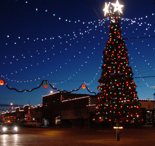A brightly lit Christmas tree adorned with colorful lights, surrounded by festive decorations in a twilight setting.