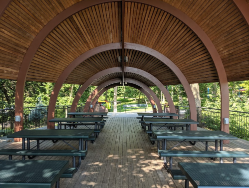 A wooden pavilion with arched beams and picnic tables, surrounded by greenery in a park setting.