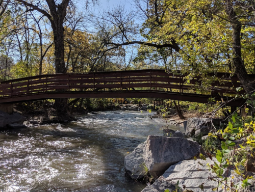 A wooden bridge spans a flowing creek, surrounded by trees and rocks under a clear blue sky.