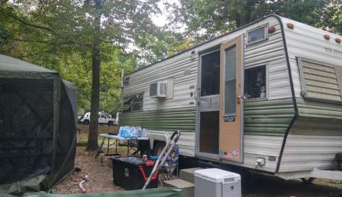 A vintage camper parked in a wooded area, with a tent and outdoor table nearby, surrounded by trees.