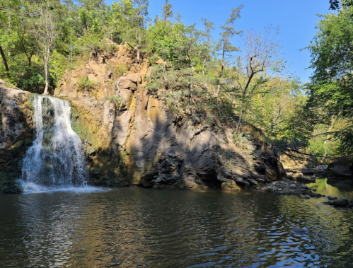 A serene waterfall cascades into a calm pool, surrounded by lush greenery and rocky terrain under a clear blue sky.