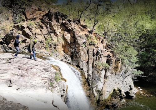 Two hikers explore a rocky area near a waterfall surrounded by trees and greenery.