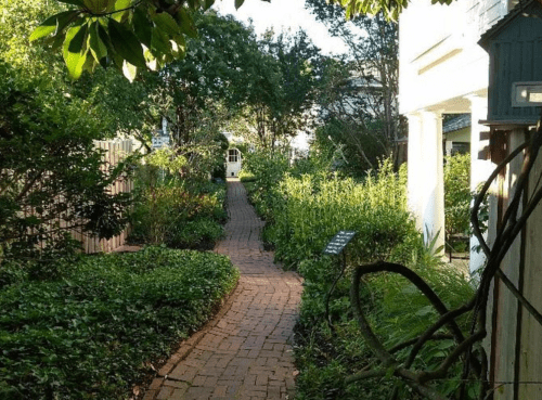 A winding brick path through a lush garden, surrounded by greenery and trees, leading to a white building in the distance.