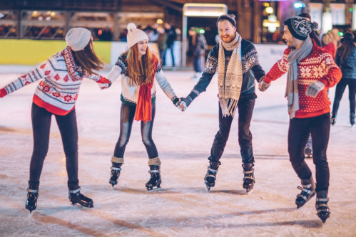 Four friends in festive sweaters skate together on an ice rink, holding hands and enjoying the winter atmosphere.