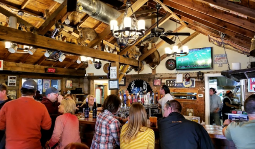 A lively bar scene with patrons socializing, rustic decor, and a TV showing a sports game in a cozy setting.