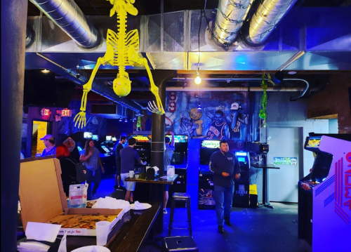 A lively arcade with vintage games, a hanging skeleton decoration, and people enjoying food and drinks.
