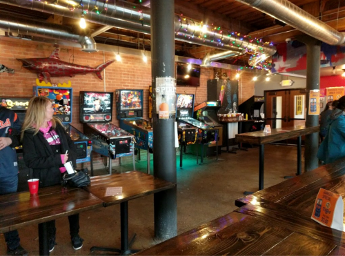 A lively arcade with pinball machines, colorful lights, and wooden tables in a casual setting.