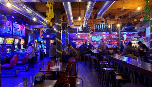 A vibrant bar with arcade games, colorful lights, and patrons enjoying drinks at wooden tables.