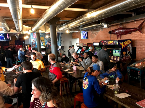 A lively arcade bar scene with people enjoying games, food, and drinks in a vibrant, retro atmosphere.