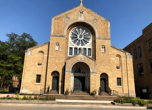 A large brick church with a circular stained glass window and a cross on top, set against a clear blue sky.