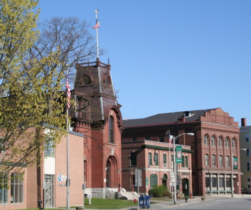 Historic brick buildings with flags, clear blue sky, and trees in the foreground.