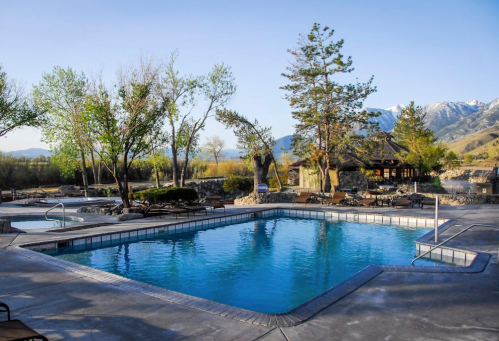 A serene outdoor pool surrounded by trees and mountains, with lounge chairs and a stone patio in the background.